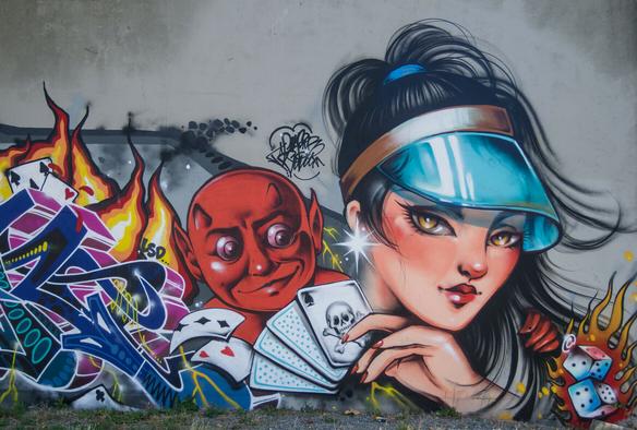 A vibrant graffiti mural depicts a stylized woman with long black hair pulled back in a ponytail, wearing a teal visor. She holds a hand of playing cards featuring a skull and crossbones.  Beside her is a cartoonish red devil, also holding playing cards. Flames and graffiti lettering, including numbers and playing card suits, surround them.  Dice are also visible within the flames. The background is a mottled grey wall.
