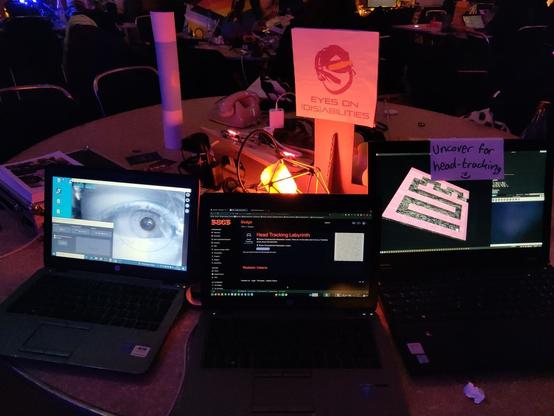 Auf einem Tisch steht ein Schild mit unserem Logo und drei Laptops. Laptop eins zeigt ein großes Auge, was digital analysiert wird. Laptop zwei zeigt Dokumentation. Laptop drei zeigt ein 3D-Labyrinth, was sich durch Kopfbewegung neigen lässt. Durch die dunkle und warme Beleuchtung, die leichte Unordnung auf dem Tisch und die Verkabelungen wirkt das Gezeigte handgemacht und persönlich.