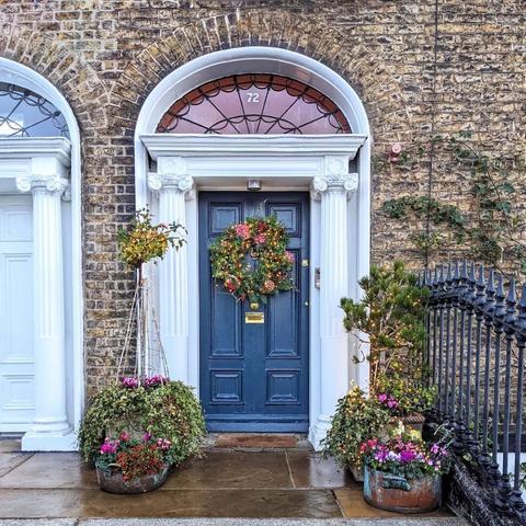 Blue Georgian door. The fanlight is lit from within wiht a purplish light. There is a wreath on the door decorated with pink and purple flowers. The wreath is covered in Christmas lights. There are potted plants on either side of the door also decorated with Christmas lights.