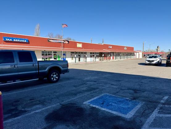 A photo taken in a parking lot. The camera is positioned at the tip left of a handicapped parking spot, looking out across the lot. A large pickup truck is parked two spots away, to the left. A white Chevrolet Malibu is parked at the end of the next row, and the illuminated brake light of a black pickup truck is just visible next to it. Across the parking lot is a row of shops with a covered walkway out front. The shops are mostly fronted by white stucco with windows, many of which have Christmas decorations, though there is a small pat h of stone wall. The roof over the walkways and a facade above it for signage is red steel. The visible signs for the shops are, from left to right, ‘TAX SERVICE’, ‘HAIR DEPOT’, something illegible, ‘PIZZA PARLOR’, and ‘C&C MOTORCYCLE 505-501-0043’. Beyond the end of the row of shops is a fast food restaurant. Their sign has a stylized hamburger with eyes and a mustache, wearing a top hat. Below, the name of the restaurant, Dandy’s Burgers, is visible, though the rest of the sign is too small to read.
