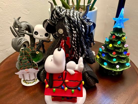 A collection of figurines on a wooden table, featuring a plush Krampus, a Funko Pop Jack Skellington, a paper pop-up Santa Claus wearing a Christmas tree hat, a Funko Pop Snoopy sleeping on his bright red doghouse adorned with Christmas lights, and a small lit ceramic Christmas tree.