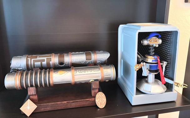 A wood stand holding two lightsaber hilts with their associated pins, next to a statuette of R-3X from Star Tours.