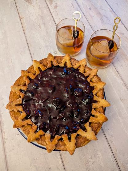 A rectangular photo of a pie, with two cocktails to its top right, on a white wooden background. The pie has a golden brown crust, decorated with little pastry fir trees all around, surrounding a dark-red cherry topping. The cocktails are two whisky-coloured drinks in rocks glasses with one large ice-cube each and a cherry garnish on a golden cocktail stick.
