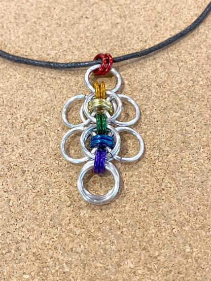 A chainmail pendant on a black cord. The pendant is made of larger, silver-coloured rings, joined together by smaller, colourful rings. The colourful rings are in the colours of the rainbow pride flag.