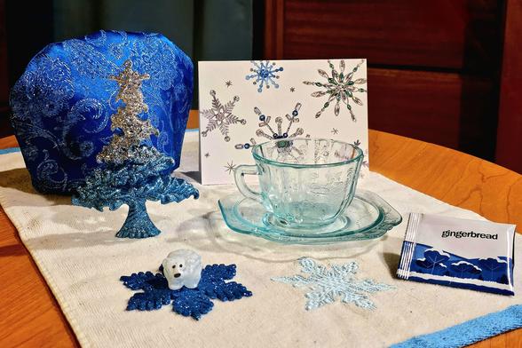 A tea tableau in chilly, icy colors of turquoise, white, silver, and deep blue. There is a sparkly fir tree, some glittery snowflakes, a quartz polar bear, a shimmering brocade tea cozy, a vintage aquamarine glass cup and saucer, and the tea is Gingerbread black tea from Adagio.