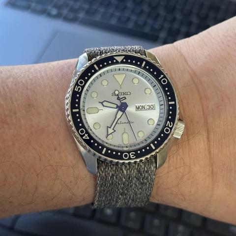 Watch with stainless steel case with brushed upper surfaces and polished sides, a 4:00 crown, an ice blue dial with day/date at 3:00, blue diver-style arrow hands and blue seconds hand, diver-style applied indices, and gray tweed-like fabric strap.