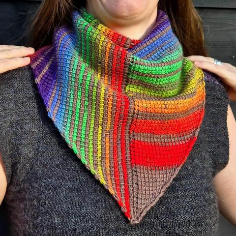 The model is standing in front of a black wall with wood designs. They are wearing a grey shirt and they have their hands on their shoulders. They are wearing the Deel Shawl wrapped around their neck so that the bottom V of the shawl is in the centre of their chest. The design is highlighted with the horizontal stitches on the right (when looking at the picture) and vertical stitches on the left (when looking at the picture). The shawl features rows of colour, rainbow colour is striped with a lovely soft brown colour.
