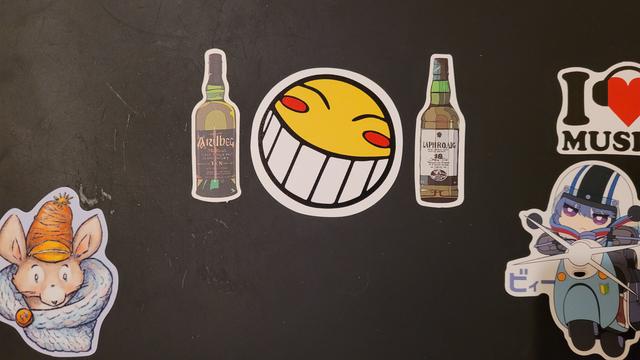 Stickers on an old Lenovo Thinkpad laptop: in the center is the smiley face Ed uses in Cowboy Bebop, on either side of that are bottles of Scotch, in the bottom left is a mouse in a cozy sweater and hat, in the bottom right is Rin from Yuru Camp on her scooter, and above her is I "heart" music (the heart has headphones around it). The rest of the laptop is blank.