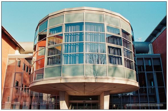 Mehrstöckige, runde, großzügig verglaste, mit pastellig hellblauen Fassadenplatten unter- und oberhalb der vier Fensterreihen versehene, auf zwei Betonpfeilern neben den Eingangstüren ruhende Konstruktion von großer architektonischer Klarheit und Geradlinigkeit.