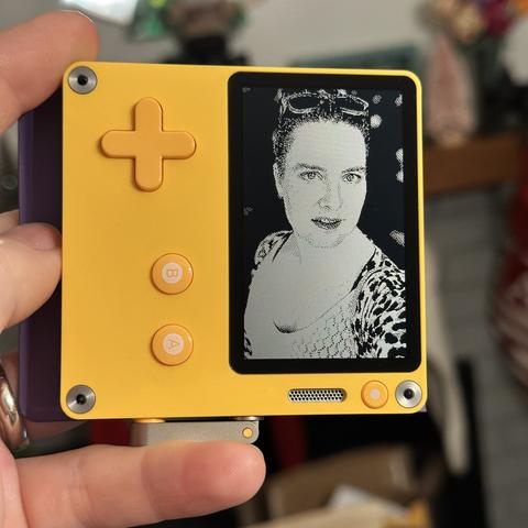 My hand holding a Playdate rotated 90° and with the screen displaying a selfie of me, a white woman wearing sunglasses on my head and a leopard print sweater. The image is dithered in black and white using the Atkinson algorithm.