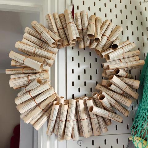 A wreath hanging halfway off an IKEA pegboard with a sea green net bag in one corner. The wreath itself consists of many rolled up book pages, gently yellowed by age, tied up around the middle with copper wire to fasten them.