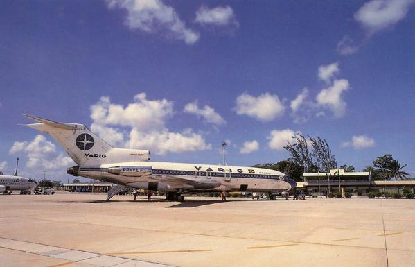 Airplane sits at the terminal under bright blue skies. There are a few cotton ball shaped clouds. Trees sway in the wind.