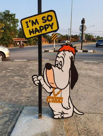 Streetart. A cartoon dog (Droopy dog) about 1.50 m tall stands as a cardboard display on a traffic island. In his right hand he holds up a sign with the inscription "I'm so happy". There by he makes a very sad face.