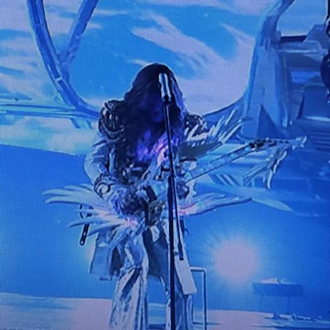 A performer playing an electric guitar on stage, dressed in a metallic, elaborate outfit. The background features bright blue lighting and dynamic visuals.
