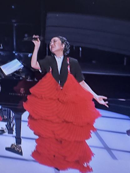 A performer wearing a striking red layered dress sings passionately on stage. She holds a microphone and expresses emotion, with a piano and musicians visible in the background.