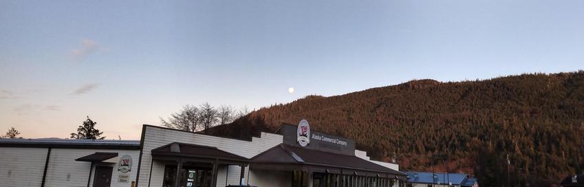 The full moon appearing in the late afternoon above the flank of a many-treed mountainside, with a rural Alaskan grocery store frontage in the foreground, the sign "Alaska Commercial Company". The sky is peach colored at horizon, shading to pale blue above, with only a few clouds.