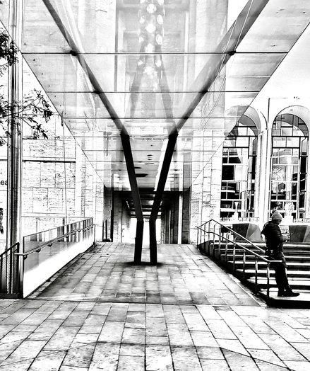 Black and white photo of the side entrance of the New York City ballet at Lincoln Center. Two dark beams are in the center of the walkway leading up to a ceiling where there is a reflection of the lights coming down on the right two of the iconic arches of Lincoln Center can be seen