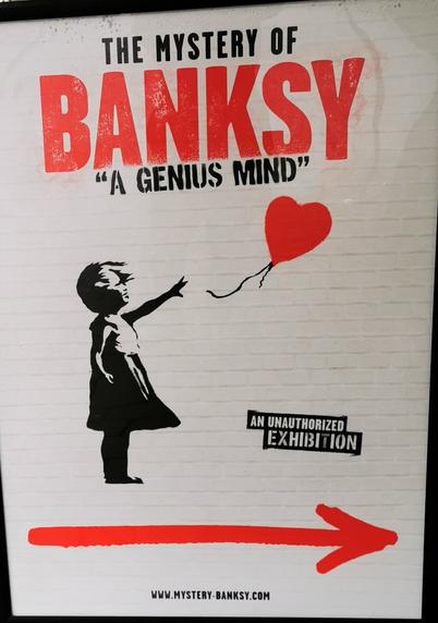 Ein Hinweisschild zeigt mit einem roten Pfeil nach rechts den Weg zur Ausstellung "The Mystery of #Banksy" mit dem Untertitel "A Genius Mind". Unter dem Titel sieht man das Bild "Girl with Balloon" und den Hinweis "An Unauthorized Exhibition". Das Bild zeigt ein Mädchen mit wehenden Haaren und Kleid, das nach einem roten, herzförmigen Ballon greift.