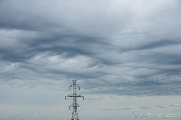 A grey threatening sky with rounded wave-like clouds looks low over a tall high-tension electrical pylon.
