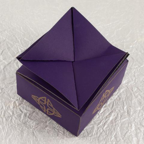 A small, squat, square, deep violet twist-top box with a Celtic knot engraved in a pale brown on each side (top-down view).