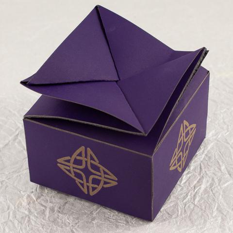 A small, squat, square, deep violet twist-top box with a Celtic knot engraved in a pale brown on each side (slightly higher angle).