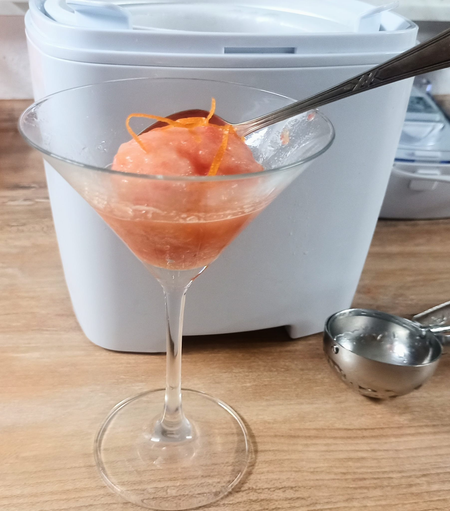 Vor der Eismaschine steht ein Martiniglas mit einer Kugel orangeroten Eises und einigen Orangenzesten. Daneben liegt ein Eisportionierer
