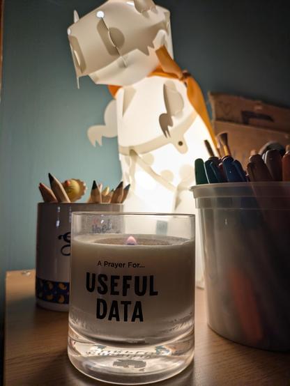 A lit candle - "A Prayer for Useful Data" in front of a lit T-Rex lamp and two containers of art supplies