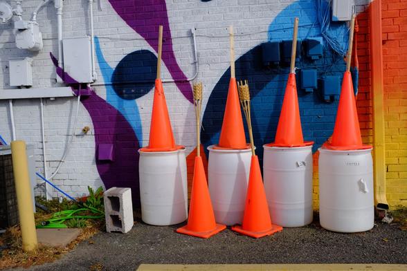 A horizontal color photograph showing a set of six bright orange traffic cones, four of which are set atop white cans. All six have some sort of wood stick poking out of their tops. They are in front of a mural painted onto the back of a commercial building. The mural is an abstract art of forms, including ovals, circles, and indeterminate organic shapes, all in purples, blacks, blues and some deep oranges and yellows; set against a white background.