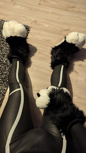 The image shows a person seated on the floor wearing a black Blue70 Bolt wetsuit with reflective white accents. They are also wearing large, plush animal-like paw coverings. The feet have white, padded, paw-like toes with black claws, and the wrists are encircled with black furry material resembling animal fur. The paws add a playful, animalistic touch to the overall appearance. The background features a light wood floor with visible grain and a black-and-white speckled rug nearby.