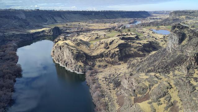 On the left side of this photo, the Snake River comes straight down afger gaving cleared a bend in the middle of a huge canyon. The sky and white clouds are mirrored in the surface. The terrain to the right of the river is rugged and rocky in a color palette of dark grqt, yellow and green. A thin strip of light-blue sky is visible at the top of the photo, and under it you can just make out a row of houses at the rim of the canyon.