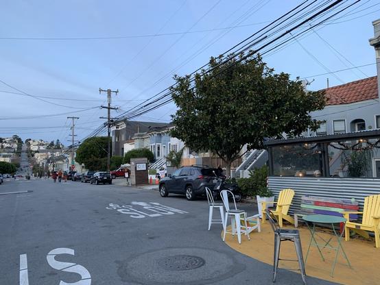A street with a painted yellow area of pavement with a bench and chairs. Further on there are groups of people walking in the street