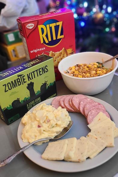 Photo of a plate of cheese, sausage and potato salad. There's a Zombie Kittens tabletop game on the left, a bowl of salad on the right, and a box of Ritz crackers behind them.