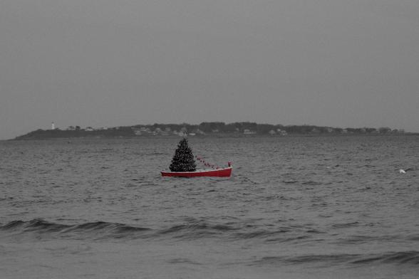 Photo of a red rowboat floating about 50 yards offshore. F/5, 1/200, 135mm, ISO 1600.   A Christmas tree sits at the boat's stern, a red-capped elf or gnome at its prow.  The photo was selectively desaturated in Darktable to feature only its red hues.  I think the land mass in the background is Baker's Island.