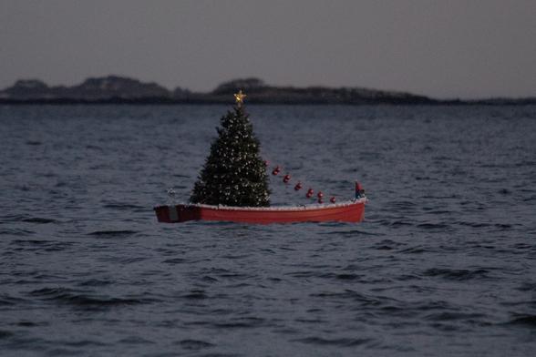 Grainy photo of a red rowboat floating about 50 yards offshore.  F/5.6, 1/160, ISO 6400, 275mm.  The boat hosts a decorated Christmas tree to its stern, and a red-capped elf or gnome at its prow. The golden star atop the tree is glowing softly.