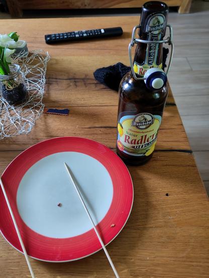 Ein leerer Teller zwei Holzstäbchen und eine Flasche alkoholfreies Radler auf einem Holztisch.