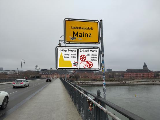 Blick von der Theodor-Heuß-Brücke auf Mainz. Im Vordergrund das Ortseingangsschild der Landeshauptstadt Mainz. Darunter ein Hinweisschild für "Heilige Messe" (mit Uhrzeiten) und rechts daneben für "Critical Mass – Jeden 1. Freitag im Monat 18.30 Uhr".