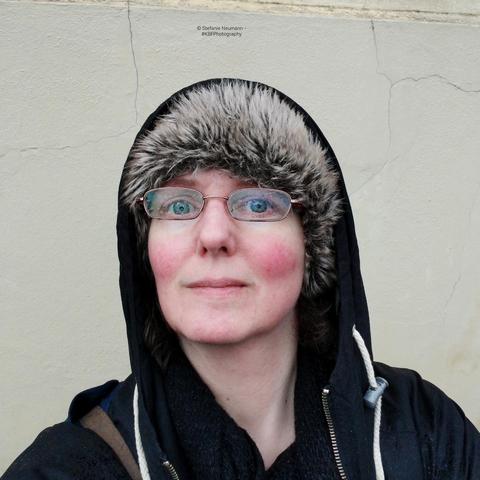 A selfie of me with hat, and hood, wet jacket, and wet glasses, in front of a yellow wall.