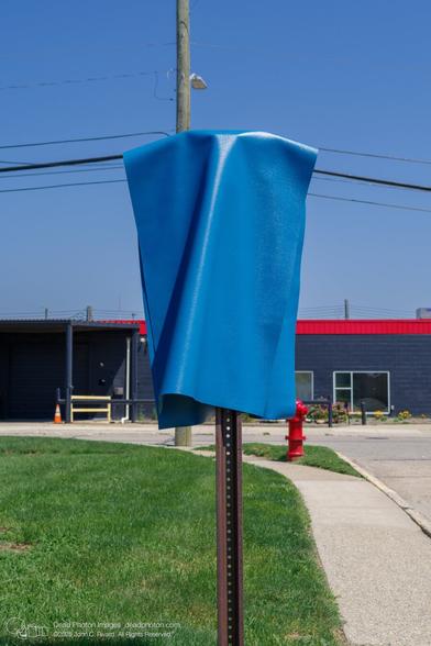 A bright blue tarp or cover is draped over a rusty metal post, likely a parking sign or similar utility post. The post is situated in a grassy area next to a sidewalk and a street, in front of a dark-gray building with a red roof.