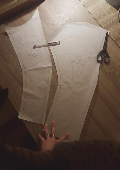 Two paper pattern pieces laid on a desk, poorly lit. They are hand drawn with some cursive markings and seam allowances drafted on. The lower sleeve, the one going under the arm, is quite narrow. The upper sleeve has a pretty wide sleeve head, not very curvy. They both have swoops and curves to follow the shape of the arm when it's bent, and to reduce bulk at the upper back sleeve. They're on the flimsy Nordic patterning paper type, which is see through, light yet durable. Due to the lightness the pieces are held down by a gel pen, a pair of scissors and Sini's hand. The lower portion of the photo is taken up by the side on a brown and white tabby cat who absolutely had to participate.