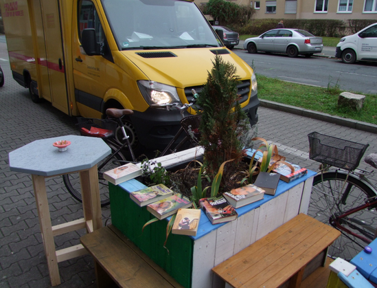 Eine bunt angemalte und bepflanzte Holzkonstruktion mit kleiner Sitzbank in der Mitte, einer seitlich angebrachten Bank sowie ein kleiner Stehtisch mit hellblauer Platte stehen am Rand einer gepflasterten Fahrbahn. Auf den Randabdeck-Brettern der Konstruktion sind Bücher ausgelegt, offenbar als Angebot zum Mitnehmen.
Daneben parkt ein Fahrrad, schräg dahinter ein Post-Lieferauto.
