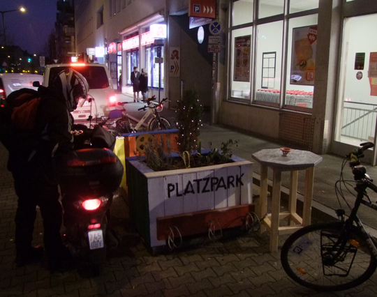 Es ist dunkel, das Licht von Schaufenstern bzw. Läden erhellt die Straße.
Eine bemalte und bepflanzte Holzkonstruktion mit kleiner Sitzbank und einem Stehtisch mit hellblauer Platte stehen am Fahrbahnrand vor einem Supermarkt. Ein Teil der Holzkonstruktion trägt die Aufschrift "PLATZPARK". Ein kleiner Winterbaum darin trägt eine leuchtende Lichterkette.
Davor parkt ein Fahrrad, schräg dahinter wird gerade ein Moped abgestellt.