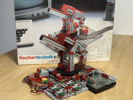 A pick and place robot made of Fischertechnik. It features a single arm that can move back and forth diagonally with an electromagnet at its tip. It can also rotate around its vertical axis. In front of the robot are various spots in different heights where metal plates can be picked and placed. The various motors, sensors and buttons are connected to a flat control unit via many colored wires.