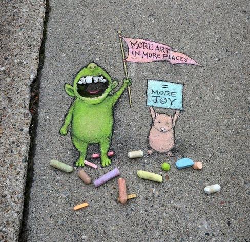 Streetart. Two small, friendly monsters with messages have been drawn in chalk on a gray sidewalk. A green monster with an open mouth stands with a banner in its hand next to a smaller mouse-like animal with a small placard in its hand. The green monster looks like a teddy bear and its banner demands: "More Art In More Places." The beige monster resembles a mouse and its placard demands: "More joy".
There are colorful pieces of chalk all around them.