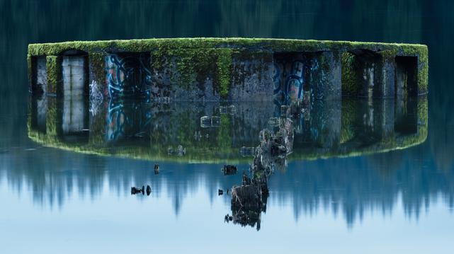 A long exposure photograph of a portion of a ruined mill in Port Moody, BC. The main portion is circular and made of concrete, with several indentations in it. This main portion has several areas with graffiti on them, and a consistent light green layer of moss covers the top. It is high tide at the time of the photo, and the ruins (as well as several old posts) are mostly submerged.