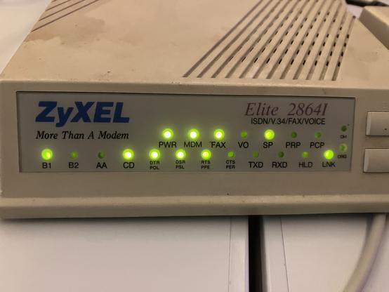 Image of a ZyXEL Elite 2864I modem with various indicator lights showing power, fax, voice, and connection statuses. The modem is beige with a textured surface and features labeled buttons on the right side.
