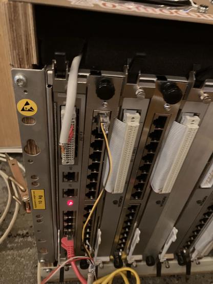 The image shows a close-up of a telecommunications or networking equipment rack. Visible components include multiple ports, indicator lights, and connected cables, with labels and warning signs present.