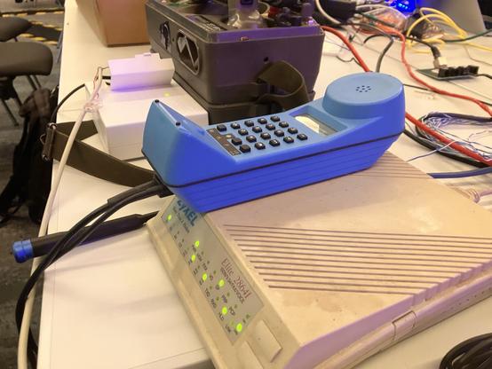 A blue vintage telephone and a white DSL modem are placed on a table alongside various cables and electronic equipment. The scene is set in a workshop or tech environment.