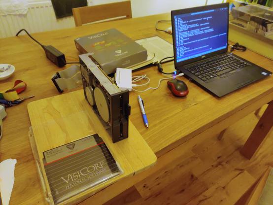 A wide shot of my kitchen table with a no-name 40-track floppy drive connected to a Greaseweazle (in the small white box at the centre of the frame) which is in turn connected to my laptop running a Debian-derived Linux distro. The original VisiCorp-branded VisiCalc disk is in the foreground at the bottom left of the frame. The lushly-presented box and manual for the VisiCalc package are in the background.