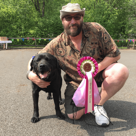 Adult black Labrador cross dog being cuddled by a kneeling middle-aged man with a beard, wearing white trainers, green shorts, a brown flowery Hawaiian shirt and a green hat, holding a Best in Show rosette and a pink goody bag. The floor is grey gravelled, and there is green foliage and trees in the background, with coloured bunting on top of a brown wooden fence.