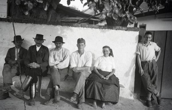 A black and white photo featuring a group of Hungarian villagers. Five are seated on a wooden bench with a whitewashed wall behind them. One is a woman in a large skirt, headscarf, and is barefooted.  The other four are men dressed in village clothes, including boots and hats. Another man is standing off to the side, leaning against the wall, with a lighted cigarette in his hand.  All six people are looking directly at the camera. In the background is what seems to be a courtyard of the house behind the wall.
